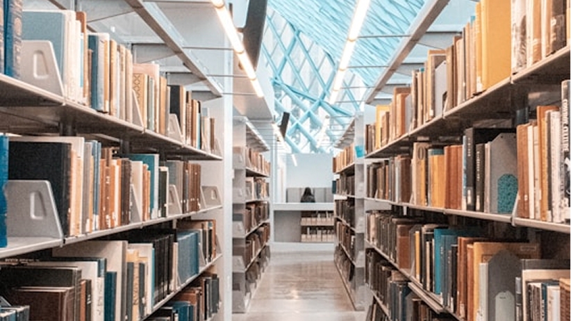 Interior of a modern library with tall bookshelves lined with colorful books on either side of an aisle, leading to a bright area with a glass, geometric skylight overhead.