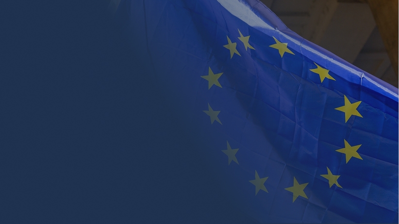 Close-up of the European Union flag rippling in the wind, showing its blue background and gold stars, with a dark blue overlay on the left side of the image.