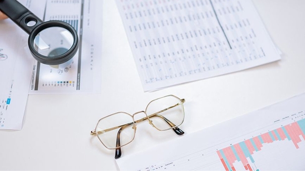 A pair of eyeglasses rests on a desk surrounded by printed charts and spreadsheets, with a magnifying glass positioned over one of the documents.