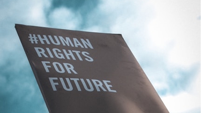 A dark banner reading "#HUMAN RIGHTS FOR FUTURE" is held up against a cloudy sky.