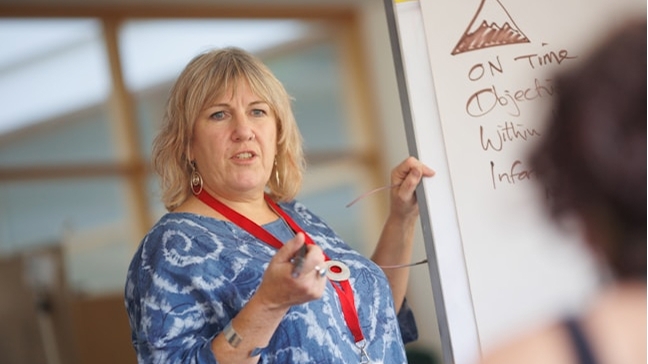 A woman wearing a lanyard gestures while speaking, standing beside a flip chart with a mountain graphic and text including "On Time" and "Objective."